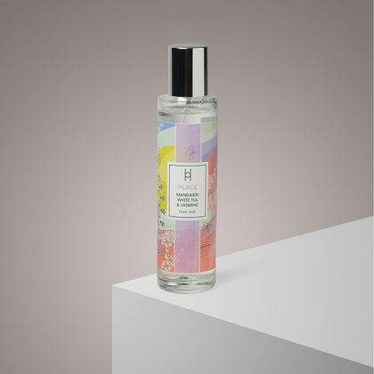 A clear glass bottle of Peace Room Mist | Lemon, Mandarin Aromatherapy Spray with a pastel label is displayed on a white platform against a gradient gray background, offering a calming scent for your space.