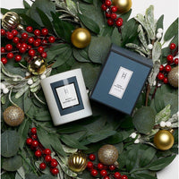 The Winter Wonderland Candle | Citrus Orange & Cedar | Limited Edition sits in its festive box on green leaves, red berries, and gold ornaments—making an elegant Christmas candle display.