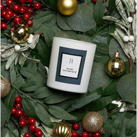 The limited edition Winter Wonderland Candle (Citrus Orange & Cedar) sits on green leaves with red berries and gold ornaments, creating a festive holiday arrangement.