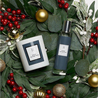 A Harrogate Organics candle and a bottle of Winter Wonderland Mist sit among green leaves, red and white berries, and gold ornaments, creating a festive display with a touch of winter fragrance.