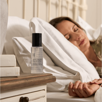 A woman sleeps peacefully in bed as a bottle of Magnesium Spray and Pillow Mist rests on the bedside table. White bedding and soft lighting enhance the tranquil atmosphere, ideal for restful sleep.