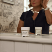 A jar of Stem Cell Lift: Hyaluronic Acid Face Cream with hibiscus stem cells sits on a bathroom counter before a mirror, reflecting both the jar and a woman in a navy blouse and silver necklace, blurred in the background.
