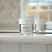 A jar of Stem Cell Lift: Hyaluronic Acid Face Cream with a white lid rests on a white bathroom surface. Infused with hibiscus stem cells, it’s shown with a blurred neutral background and sunlight streaming through a window.