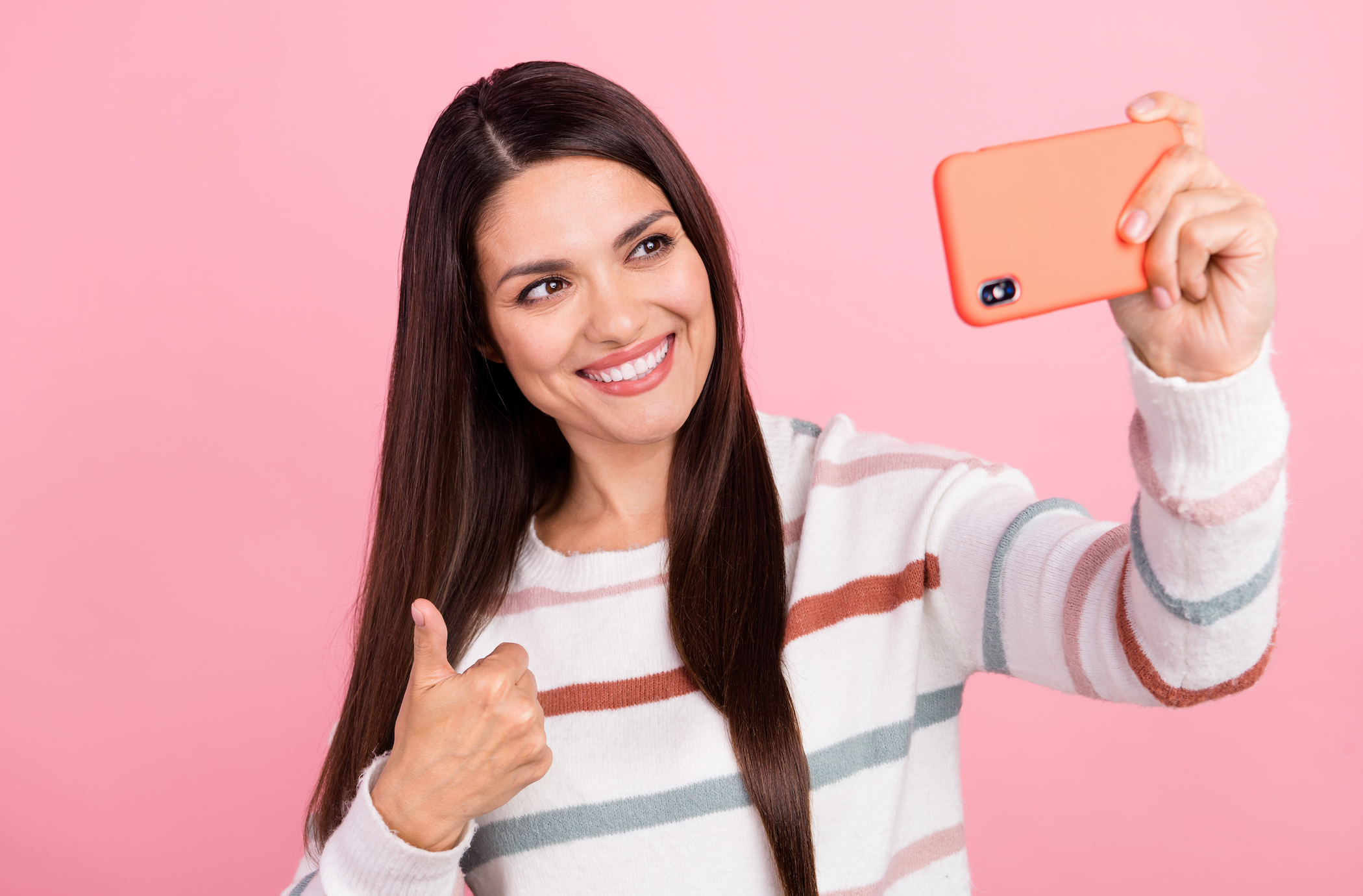 A smiling woman with long brown hair in a striped sweater takes a selfie with a coral-colored smartphone and gives a thumbs-up gesture against a pink background.