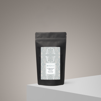A black resealable pouch with a patterned white label stands upright on a light gray surface against a beige gradient background. The label introduces Soothing Sea Kelp And Green Clay Spa Salts for a minimalist, modern spa experience.