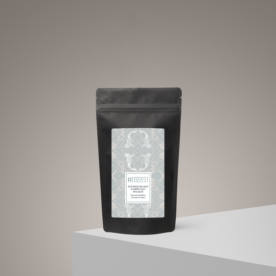A black resealable pouch with a patterned white label stands upright on a light gray surface against a beige gradient background. The label introduces Soothing Sea Kelp And Green Clay Spa Salts for a minimalist, modern spa experience.
