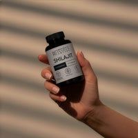 A hand with manicured nails holds a black bottle labeled Shilajit Supplement 1000mg Per Serving - 30 Day Supply - 60 Capsules against a background with diagonal light and shadow stripes.