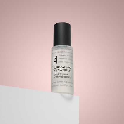 A clear bottle of Sleep Calming Pillow Spray with a black cap sits on a white surface against a pink gradient background. Its minimalist label design highlights it as an ideal sleep aid for deep rest.