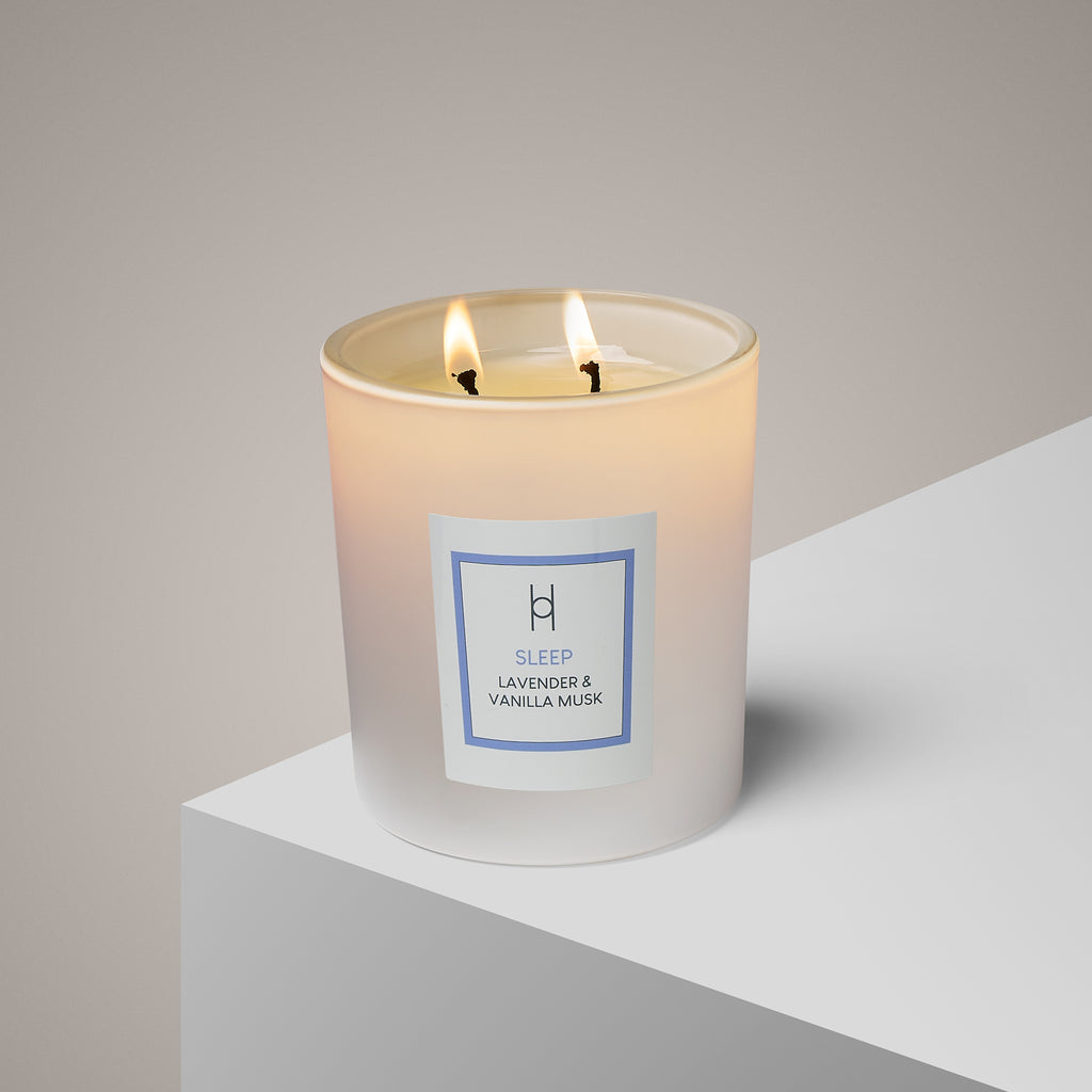 A lit Sleep Candle with two wicks rests on a white surface. Its light blue label reads “Sleep, Lavender & Vanilla Musk,” evoking calming vanilla for relaxation. The background features a smooth, neutral beige tone.