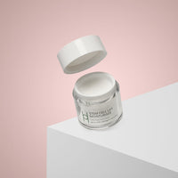 A jar from the Ultimate Women's Skincare Set | Complete 6-Piece Collection rests on a white surface with a soft pink background, its lid floating above. The mostly white jar with green and grey text promises radiant skin.