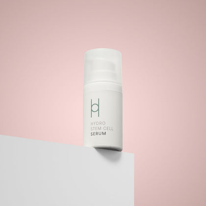 A white bottle of Hydro Stem Cell Serum rests on a white surface with a soft pink background. Infused with hibiscus extract, this anti-ageing serum helps reveal refreshed, youthful-looking skin.