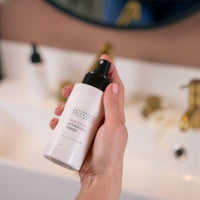 A hand holds a white bottle labeled Skin Calm Hydrating Toner from the Ultimate Women's Skincare Set | Complete 6-Piece Collection, in front of a bathroom sink with gold fixtures and a blurred mirror in the background.
