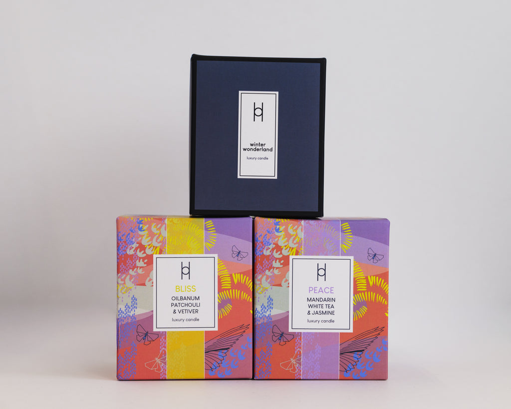 Three luxury boxes from the Trio Candle Selection are stacked: the top is plain dark blue, while BLISS and PEACE below display vibrant abstract floral designs, capturing the indulgence of luxury scented candles inside.