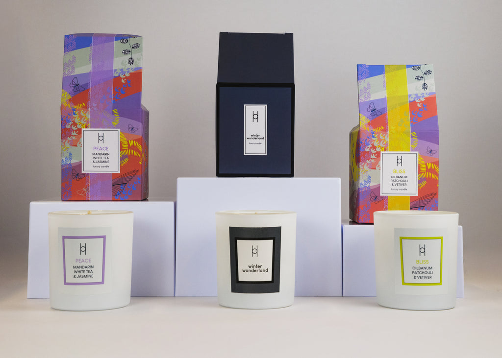 The Trio Candle Selection showcases three luxury white-jar candles, each uniquely labeled, artfully arranged in front of vibrant patterned boxes and a black box on white platforms against a neutral backdrop.