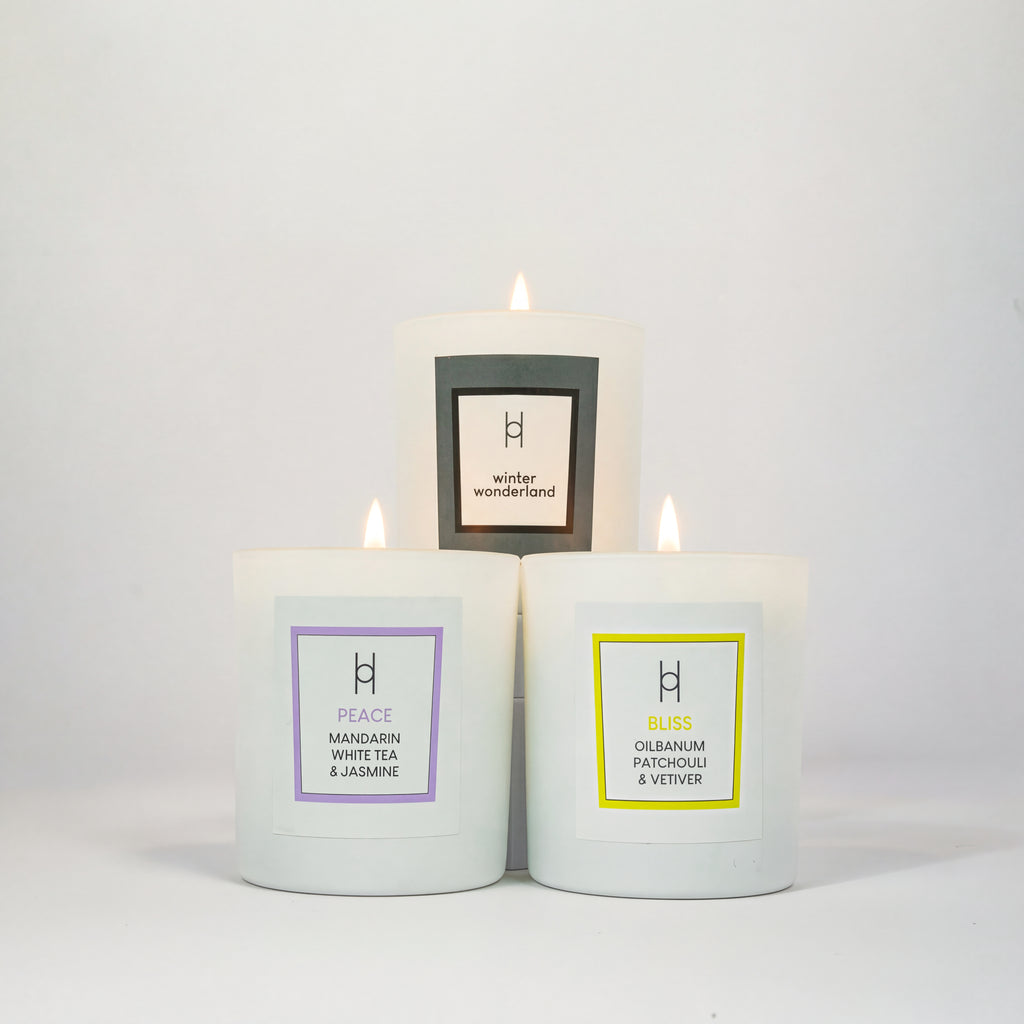 The Trio Candle Selection features three lit white luxury candles in glass containers, each labeled with a unique scent: Winter Wonderland, Peace—Mandarin White Tea & Jasmine, and Bliss—Olibanum Patchouli & Vetiver.