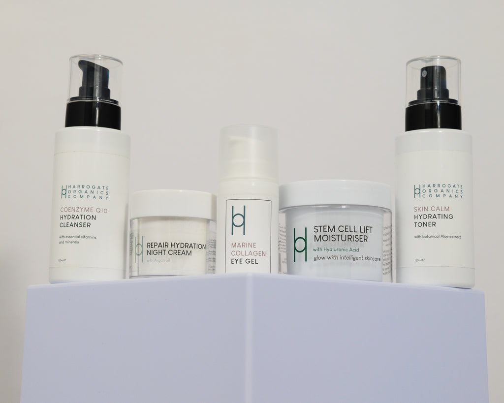 Five white bottles and jars with minimalist labels from Harrogate Organics Company make up the Ultimate Men's Skincare Routine—cleanser, night cream, eye gel, moisturizer, and toner—displayed on a white pedestal to promote healthy skin.