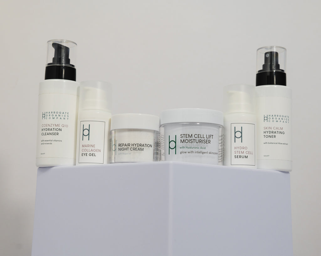 Six white containers with minimalist labels sit on a white platform. The Ultimate Women’s Skincare Routine includes cleanser, eye gel, night cream, moisturiser, serum, and toner for radiant skin.