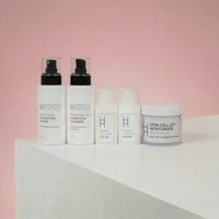 The Ultimate Women’s Skincare Set | Complete 6-Piece Collection features white bottles and jars with black pumps and minimalist labels, arranged on a light surface against a soft pink background.