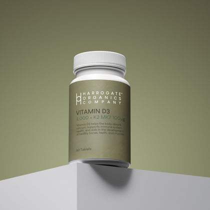 A bottle of Vitamin D3 & Vitamin K2 MK7 (4,000 IU D3 + 100µg K2) sits on a white surface against a green background. The label highlights benefits for bones, teeth, muscles, and immune support.