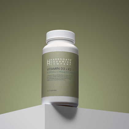 A white bottle labeled Harrogate Organics Company Vitamin D3 & Vitamin K2 MK7, a premium supplement for immune support, sits on a geometric surface with a neutral background. The label highlights 4,000 IU D3 and 110 μg Vitamin K2 per serving and contains 60 capsules.