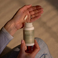 A person holds a bottle of Vitamin D3 & Vitamin K2 MK7 supplements in one hand and two capsules in the other, seated on a light surface in natural lighting—highlighting this premium Vitamin D3 & K2 MK7 supplement designed for immune support.