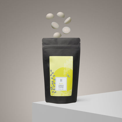 A black pouch labeled “Bliss Luxury Wax Melts | Ylang Ylang & Olibanum | 20 Melts” stands on a white surface, with five white, round aromatherapy wax melts floating above it against a neutral background.