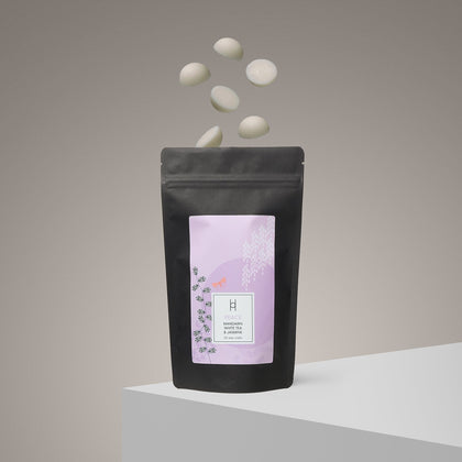 A black resealable pouch with a pastel pink label stands on a white surface, featuring Peace Luxury Wax Melts in Mandarin & Jasmine. Twenty white melts float above, evoking inner peace and a calming scent against a neutral background.