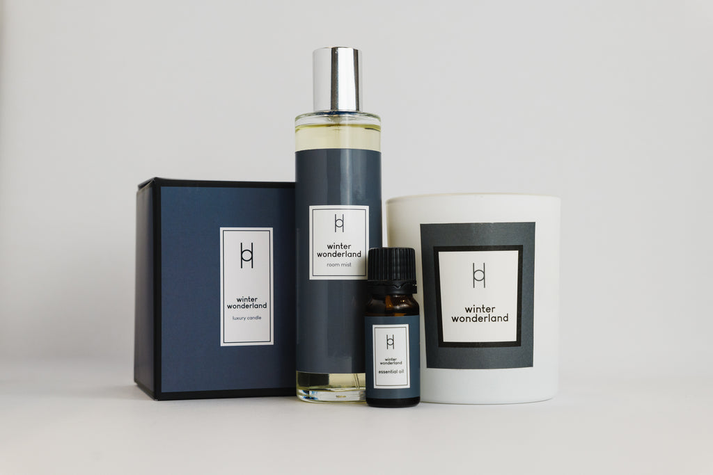 The Deluxe Home Collection - Winter Wonderland features a blue box, spray bottle, essential oil blend, and hand-poured candle—all with minimalist labels on a plain background.