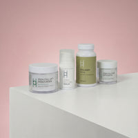 The Women's Skin Reset Gift Set | 4-Piece Anti-Ageing Collection is displayed on a white surface against a pink background, featuring minimalist packaging for the serum, collagen supplement, stem cell lift moisturizer, and face exfoliator.