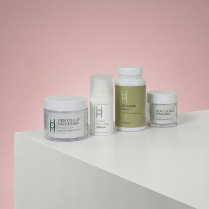 The Women's Skin Reset Gift Set | 4-Piece Anti-Ageing Collection is displayed on a white surface against a pink background, featuring minimalist packaging for the serum, collagen supplement, stem cell lift moisturizer, and face exfoliator.