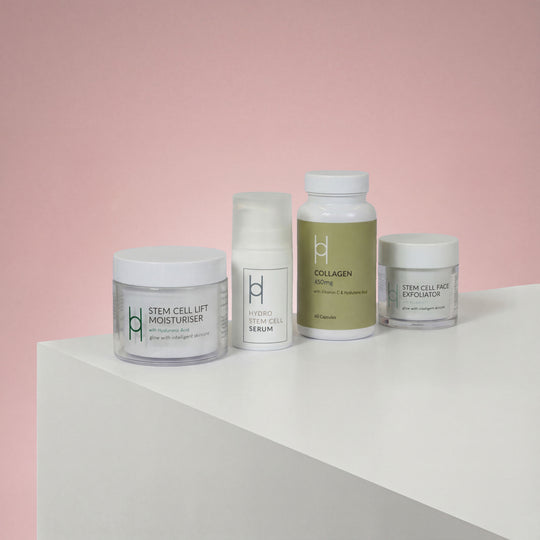 The Women's Skin Reset Gift Set | 4-Piece Anti-Ageing Collection is displayed on a white surface against a pink background, featuring minimalist packaging for the serum, collagen supplement, stem cell lift moisturizer, and face exfoliator.