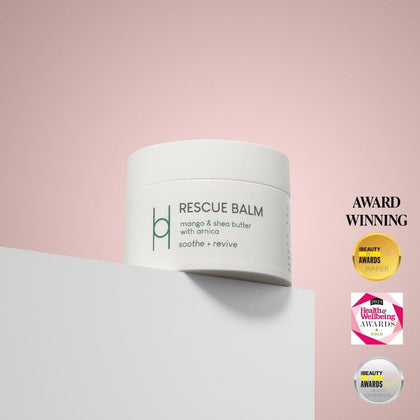 A white jar of Rescue Balm - Mango & Shea Butter with Arnica sits on a white surface against a pink background. Three award badges and "AWARD WINNING" text appear on the right, showcasing its muscle pain relief benefits.