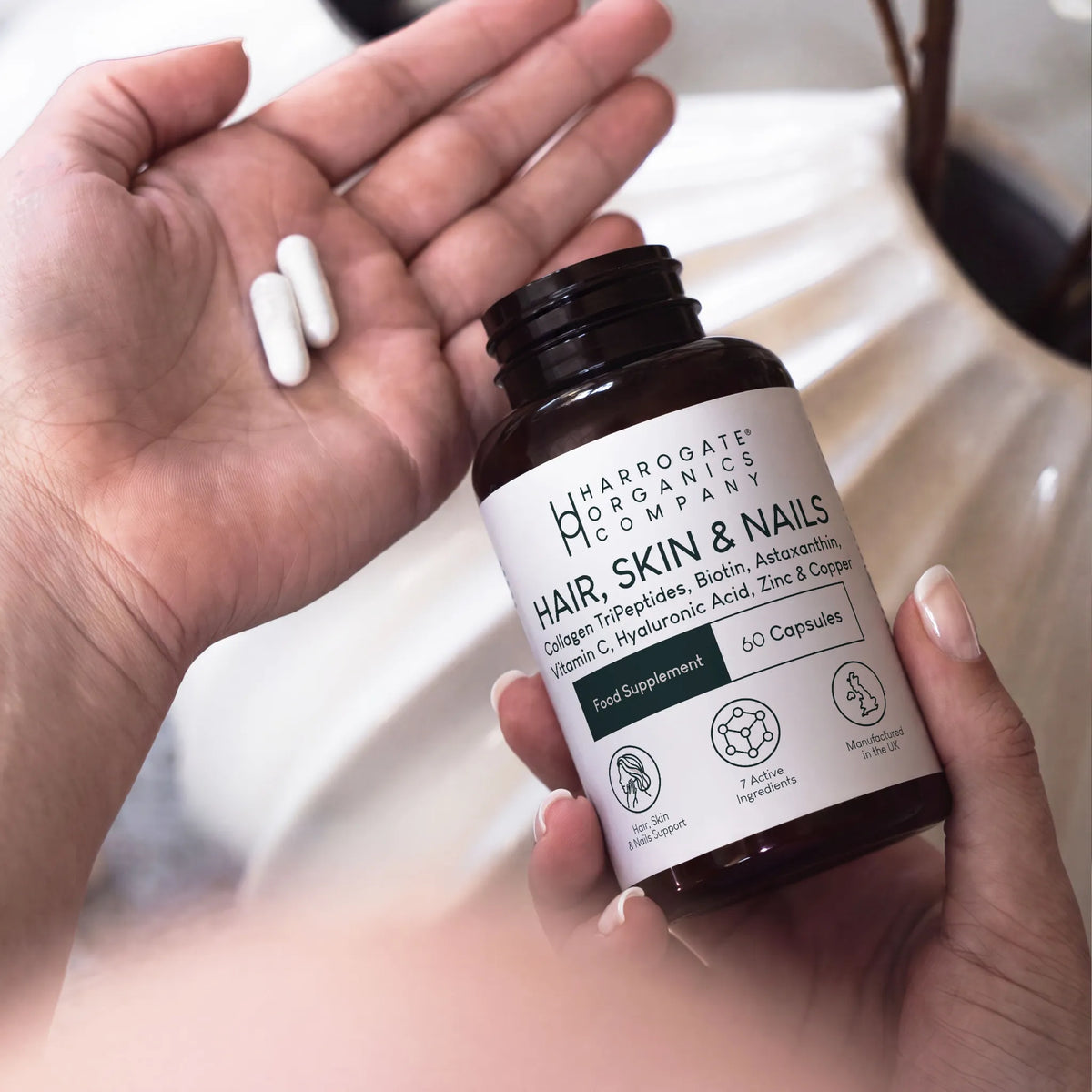 A person holds a bottle labeled Hair, Skin & Nails Complex with the lid off in one hand and two white capsules in the other. The label lists ingredients such as biotin, zinc, marine collagen, and hyaluronic acid.