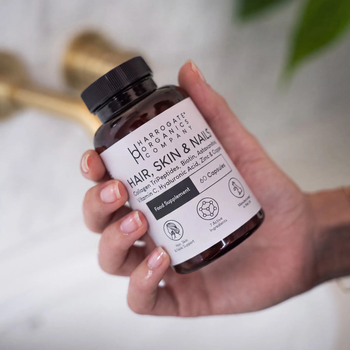 A hand holding a bottle labeled Hair, Skin & Nails Complex from Harrogate Organics Company, a food supplement enriched with Biotin and Marine Collagen for beauty support, containing 60 capsules, set against a bathroom background.
