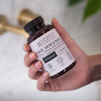 A hand holding a bottle labeled Hair, Skin & Nails Complex from Harrogate Organics Company, a food supplement enriched with Biotin and Marine Collagen for beauty support, containing 60 capsules, set against a bathroom background.