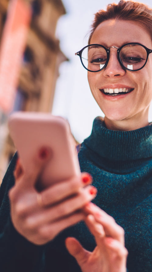 A smiling woman wearing glasses and a green turtleneck sweater looks at her smartphone outdoors on a sunny day.