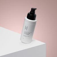 A white bottle of Coenzyme Q10 Hydration Cleanser with a black pump sits on a white surface and soft pink background, promoting collagen production for younger-looking skin.