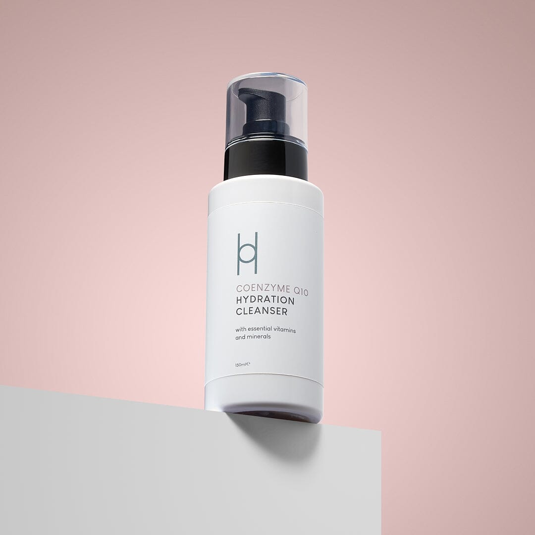 A white bottle of Coenzyme Q10 Hydration Cleanser with a black pump rests on a white surface against a soft pink background. Its label features essential vitamins, minerals, and collagen support for youthful-looking skin.