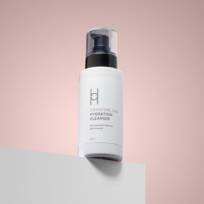 A white bottle of Coenzyme Q10 Hydration Cleanser with a black pump rests on a white surface against a soft pink background. Its label features essential vitamins, minerals, and collagen support for youthful-looking skin.