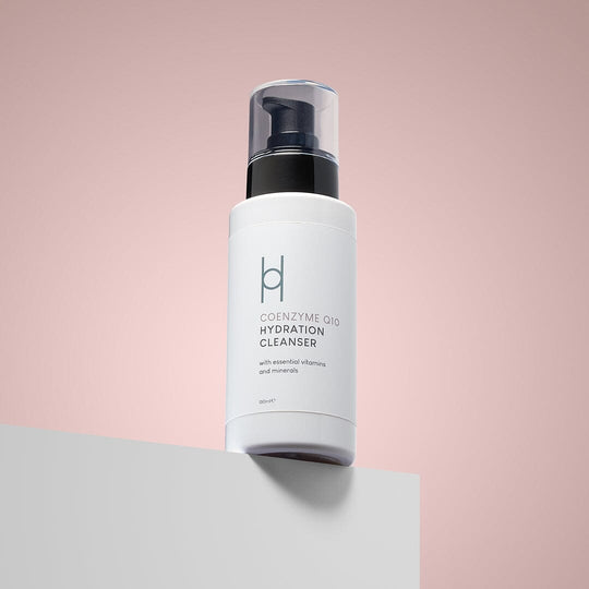 A white bottle of Coenzyme Q10 Hydration Cleanser with a black pump rests on a white surface against a soft pink background. Its label features essential vitamins, minerals, and collagen support for youthful-looking skin.