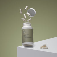 A white bottle labeled Marine Collagen 450mg with Vitamin C & Hyaluronic Acid (60 Capsules, 60 Day Supply) sits on a white geometric surface against an olive green background, with capsules flying out of the open lid and some resting beside it.