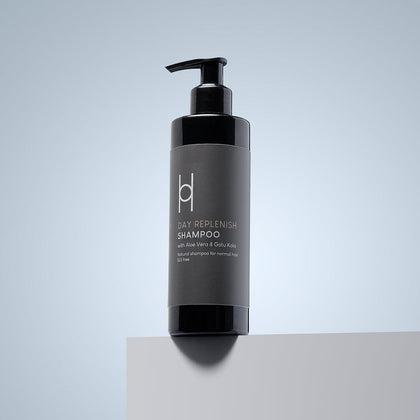 A black pump bottle of Day Replenish Natural Shampoo - With Aloe Vera & Gotu Kola - 250ml rests on a light gray surface with a soft blue-gray background, highlighting its all-natural formula for healthy hair.