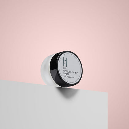 The Lip Conditioning Balm with Peppermint, featuring a black lid and minimalist label, sits on a white surface against a soft pink background—perfect for soothing chapped lips with its natural formula.