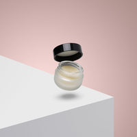 A small, frosted glass jar with a black lid floats above a white surface against a pink gradient background. The jar holds Lip Conditioning Balm with Peppermint, ideal for soothing chapped lips with nourishing cocoa butter.
