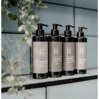 Four black pump bottles of sulfate-free shampoo and Moisture Protect Conditioner with pomegranate seed and baobab oils (250ml) are lined up on a glass shelf in a tiled shower, green leaves nearby. Grey labels with white text create a modern, clean look.