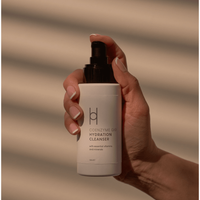 A hand with neatly manicured nails holds a white bottle from the Anti-Ageing Skincare Routine | 3-Step Daily Skincare Set, an essential for youthful skin, against a softly lit, striped background.