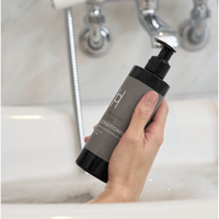 A hand holds a black bottle of Enriching Repair Conditioner with Coconut & Marula Oils (250ml) and pump dispenser over a bubbly white bathtub; the sulphate-free formula is shown near a silver faucet and shower hose.