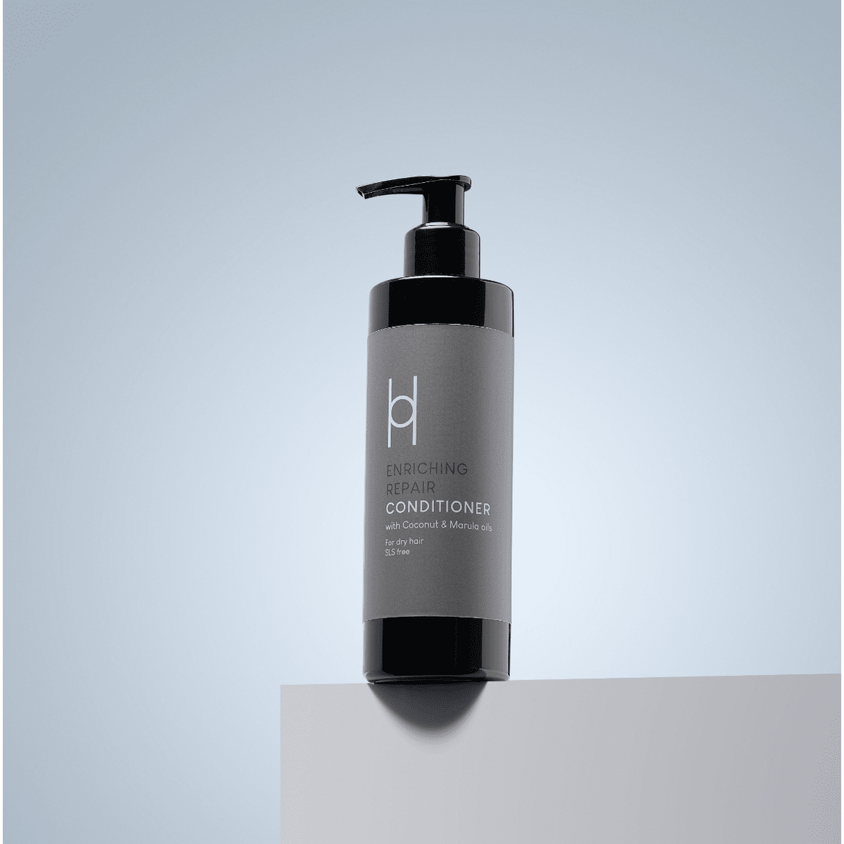 A black pump bottle of Enriching Repair Conditioner - With Coconut & Marula Oils - 250ml sits on a light gray surface against a gradient gray-blue background—your sulphate-free, revitalising hair conditioner.