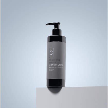 A black pump bottle of Enriching Repair Conditioner - With Coconut & Marula Oils - 250ml sits on a light gray surface against a gradient gray-blue background—your sulphate-free, revitalising hair conditioner.
