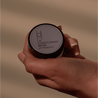 A hand holds a small round container of Lip Conditioning Balm with Peppermint, featuring a minimalist soft gray label—ideal for soothing chapped lips with nourishing cocoa butter.